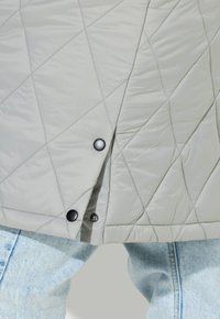 Gesteppte graue Jacke mit einem Rautenmuster, verschlossen mit schwarzen Druckknöpfen, kombiniert mit hellblauen Jeans aus Denim.