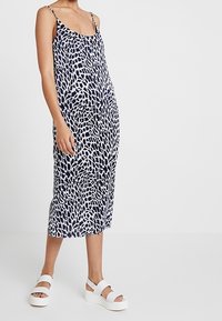 Granatowa i biała bezrękawnikowa sukienka z abstrakcyjnym wzorem, sięgająca do połowy łydki, z lekkiego materiału, w połączeniu z masywnymi białymi sandałami.