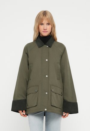 Femme portant une veste oversize vert olive avec col et poignets en velours côtelé noir, associée à un jean bleu clair, debout devant un fond blanc.