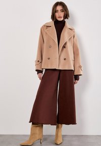 Donna in piedi, che indossa un cappotto beige doppiopetto sopra un dolcevita marrone scuro, pantaloni larghi marroni e stivali con tacco beige, davanti a una parete bianca.