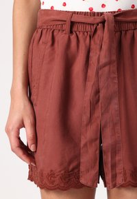 Pantaloni corti marroni con una vita alta cinturata, caratterizzati da un orlo scallop e dettagli ricamati intricati nella parte inferiore.