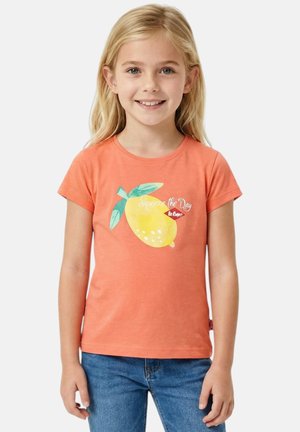 Fille souriante aux cheveux blonds portant un t-shirt orange avec un grand citron et du texte, assorti à un jean bleu, sur fond blanc.