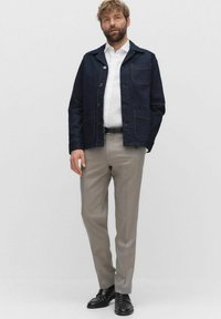 Mörkblå denimjacka med fyra fickor, bärs över en vit skjorta. Ljusgrå byxor och svarta skor fulländar outfiten.