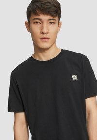 Jonge man met kort donker haar, gekleed in een zwart T-shirt met een klein wit "s.Ol" logo op de linkerborst, tegen een effen achtergrond.