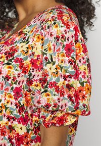 Blouse fleurie à manches bouffantes, avec un imprimé coloré de fleurs rouges, roses, orange et jaunes sur un fond clair. Tissu texturé.