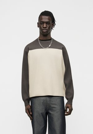 Young black man wearing a two-tone cream and brown long-sleeve shirt, dark jeans, and a silver chain necklace, standing against a plain background.