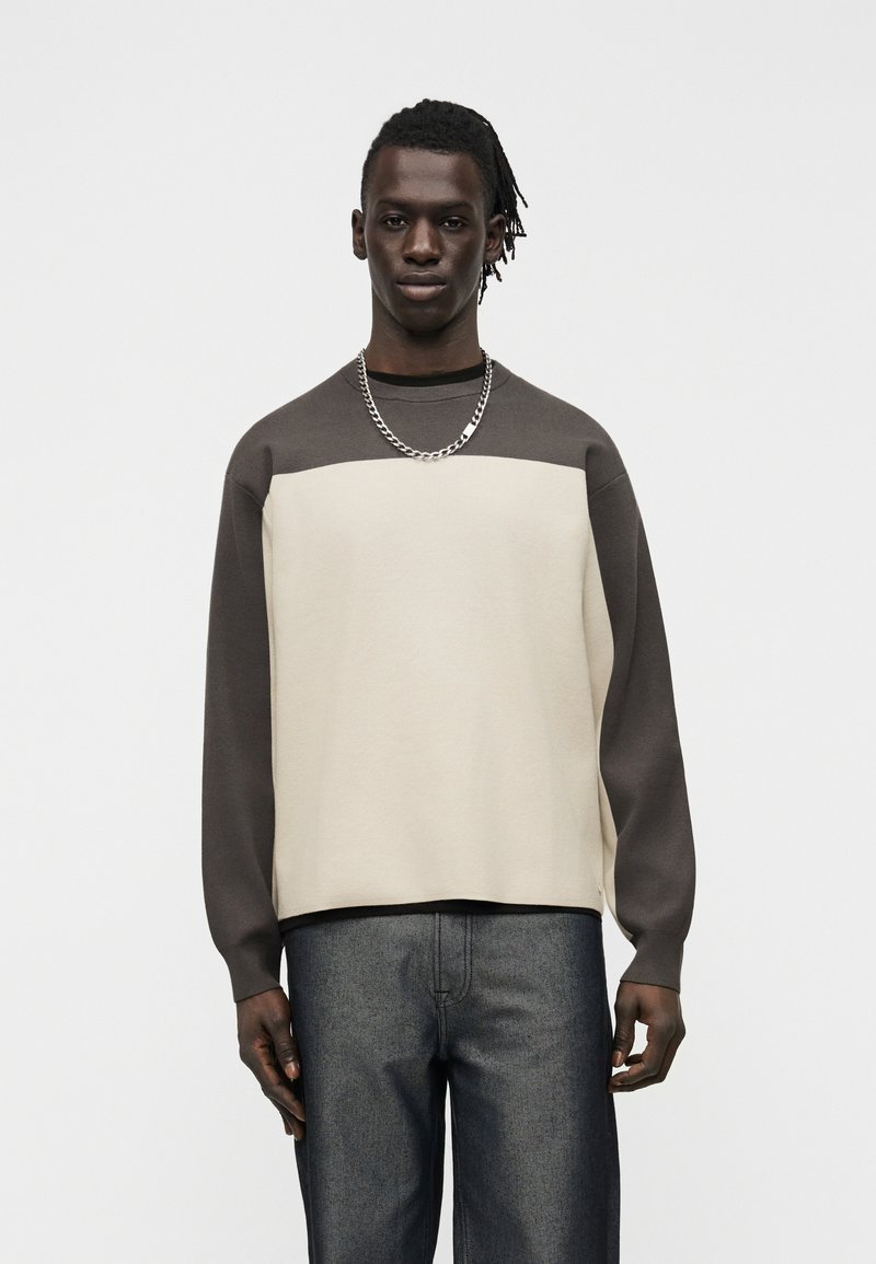 Jeune homme noir portant une chemise à manches longues bicolore crème et marron, un jean foncé et un collier en chaîne argentée, debout devant un fond uni.