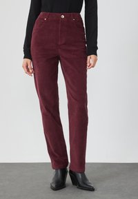 Bordeaux corduroy broek met een hoge taille, twee voorzakken en een gouden knoopdetail, gecombineerd met zwarte hoge hakken.