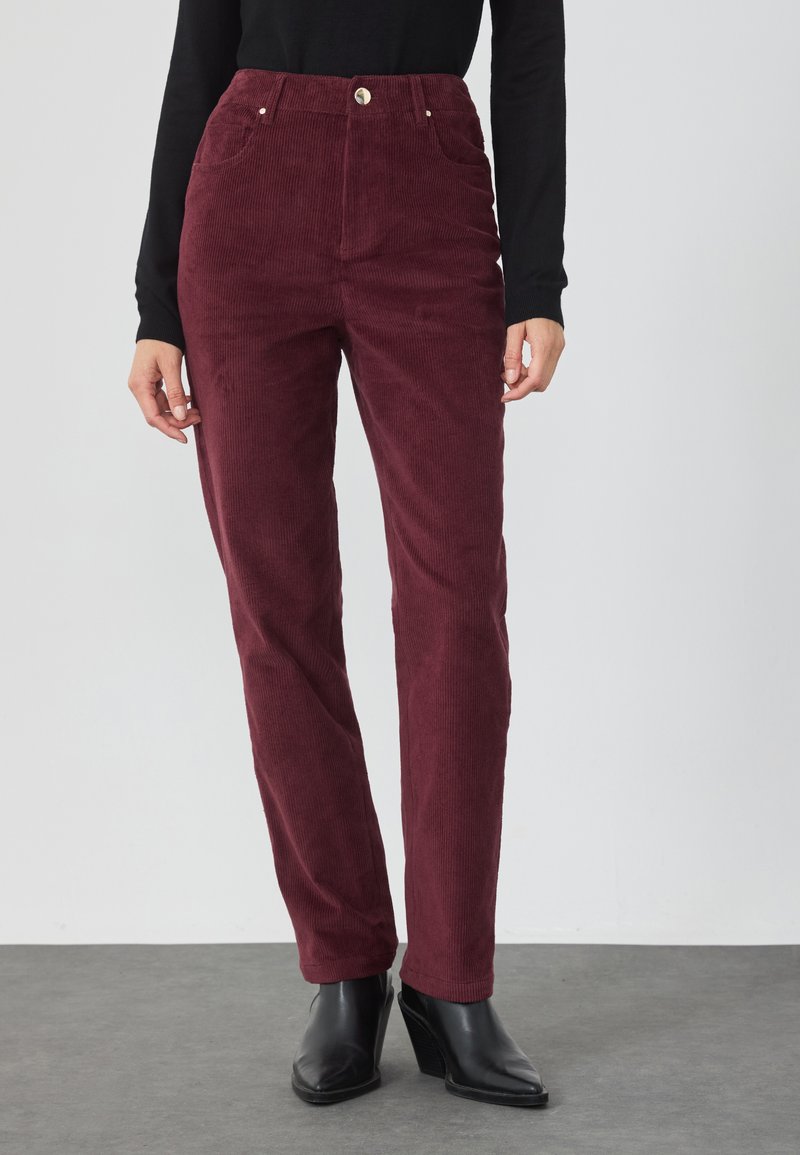 Bordeaux corduroy broek met een hoge taille, twee voorzakken en een gouden knoopdetail, gecombineerd met zwarte hoge hakken.