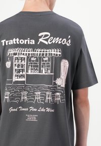 Dunkelgraues T-Shirt mit einer pinken Illustration einer Café-Szene, die ein gestreiftes Markise, Hocker und den Schriftzug "Trattoria Remo’s" zeigt.