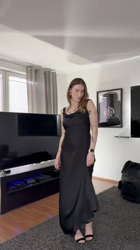 Crna satenska haljina s padajućim ovratnikom, tijesnim korzetom i lepršavom suknjom. Nošena s crnim sandalama s remenčićima; mekana tekstura i elegantan oblik.