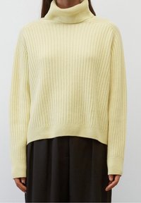 Maglione a collo alto giallo a costine con maniche lunghe; texture morbida, vestibilità ampia e orlo leggermente accorciato. Indossato sopra un indumento nero.