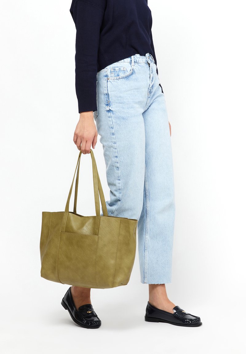Olivovo zelená taška vyrobená z texturovanej kože, so štyrmi pevnými popruhmi a priestranným interiérom, v kombinácii s svetlými džínsy a čiernymi mokasínami.