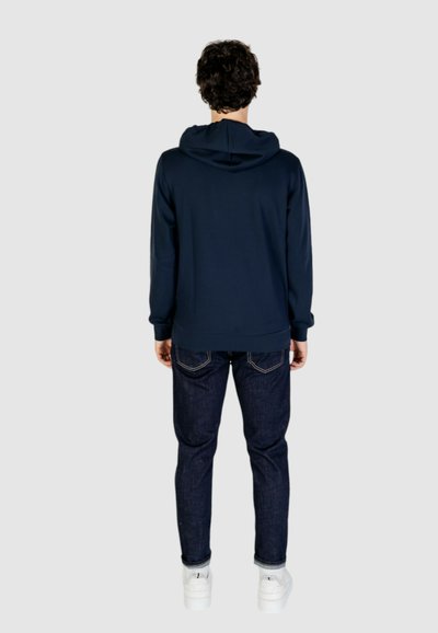 Felpa navy con cappuccio regolabile, maniche lunghe e vestibilità comoda, abbinata a jeans scuri e sneaker bianche. La texture appare liscia.