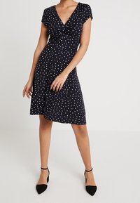 Robe noire à pois blancs avec un col en V, manches courtes style capuchon et détail torsadé sur le devant. Silhouette fluide, longueur au genou, avec une texture de tissu lisse.