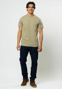 Kaki bomull t-skjorte med rund hals, korte ermer og en liten grønn logo på venstre bryst, kombinert med mørkejeans og brune støvler.