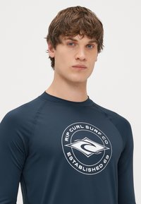 Ναυτικό μπλε rashguard φτιαγμένο από ελαστικό ύφασμα με κυκλικό λευκό λογότυπο που περιλαμβάνει το κείμενο "Rip Curl Surf Co" και "Established 69" στο κέντρο.