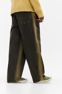 Persoană purtând blugi largi de culoare închisă, adidași verzi și un pulover galben, mergând pe fond alb simplu.