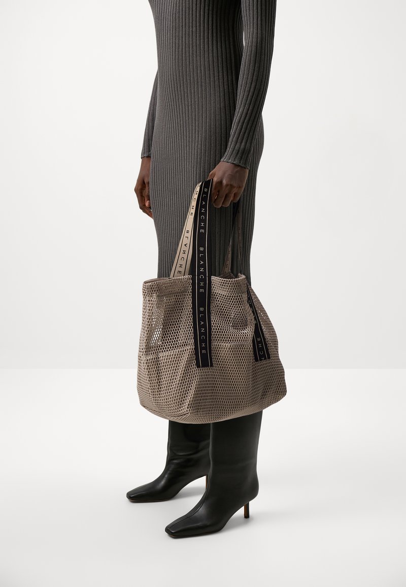Beige Netz-Shopper mit zwei schwarzen, bedruckten Trägern, strukturierter Form und offenem Design; Modell trägt schwarze knielange Stiefel.