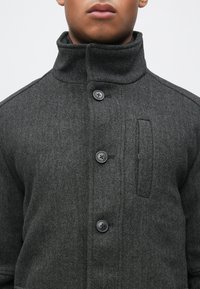 Siv pleteni plašč z ribjo kostjo z visokim ovratnikom, opremljen s črnimi gumbi in žeparjem z našitkom. Material je teksturiran in nudi strukturiran videz.