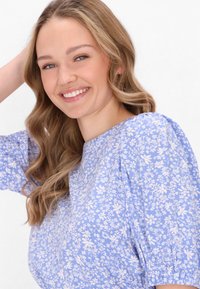 Hellbluse mit floralen Mustern in Weiß, mit Puffärmeln und einem runden Ausschnitt. Die Haare in sanften Wellen frisiert, mit einem lächelnden Gesichtsausdruck.
