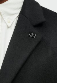 Sort filt blazer stof med en stor, struktureret krave. Sølv rektangulær nål med sorte accenter fastgjort til klappen. Hvid skjorte under.