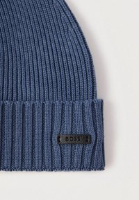 Căciulă albastră tricotată, având o textură reiată și o etichetă de piele neagră cu logo-ul "BOSS" pe marginea întoarsă.