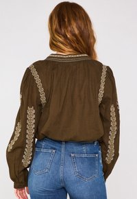 Blusa verde oliva con pieghe e decorazioni ricamate sulle maniche, abbinata a jeans blu. Il tessuto ha una texture morbida.