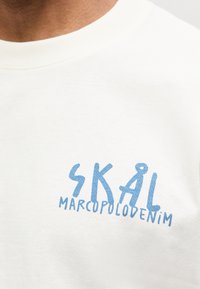 Κοντινή λήψη λευκού μπλουζάκι με μπλε κείμενο "SKAL" και μικρότερο κείμενο "MARCOPOLODENIM" τυπωμένα στο στήθος.