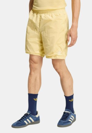 Uomo con pantaloncini giallo chiaro, calzini Adidas blu navy e scarpe da ginnastica Adidas blu, in piedi davanti a uno sfondo semplice.
