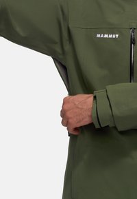 Personne ajustant la ventilation zippée sous le bras sur une manche de veste Mammut verte avec le logo de la marque visible sur la poitrine.