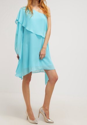 Kobieta ubrana w bezrękawnikową, asymetryczną sukienkę w kolorze aqua oraz srebrne wysokie obcasy, stojąca z jedną lekko ugiętą nogą na jednolitym tle.