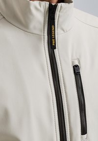 Light gray jacket with a high collar, featuring a black zipper and a branded pull tab. Texture appears smooth with a fitted design.