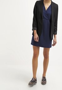 Marinblå klänning med V-ringning, lager under en svart blazer med upprullade ärmar, i kombination med mörka, mönstrade skor med metalliska detaljer.