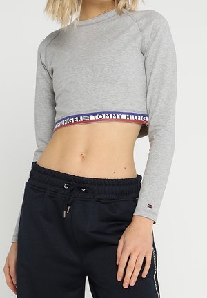 Vrouw draagt een grijs cropped langermig shirt met een Tommy Hilfiger logoband en zwarte sweatpants met trekkoord, hand rustend op dij.