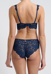 Námořnická krajková souprava spodního prádla s květinovými vzory, vlnitými okraji a nastavitelnými ramínky. Zadní pohled ukazuje složitý krajkový design a háčky.