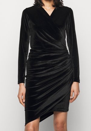 Femme portant une robe portefeuille en velours noir à manches longues avec des détails froncés et un ourlet asymétrique sur un fond clair uni.