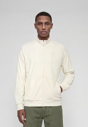 Homme portant une veste écrue zippée à col montant et un pantalon vert, debout avec une main dans la poche de la veste sur un fond uni.