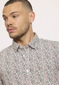 Chemise à boutons avec un motif abstrait multicolore en vert, rouge et blanc. Présente un col classique et des boutons blancs. En matériau coton.