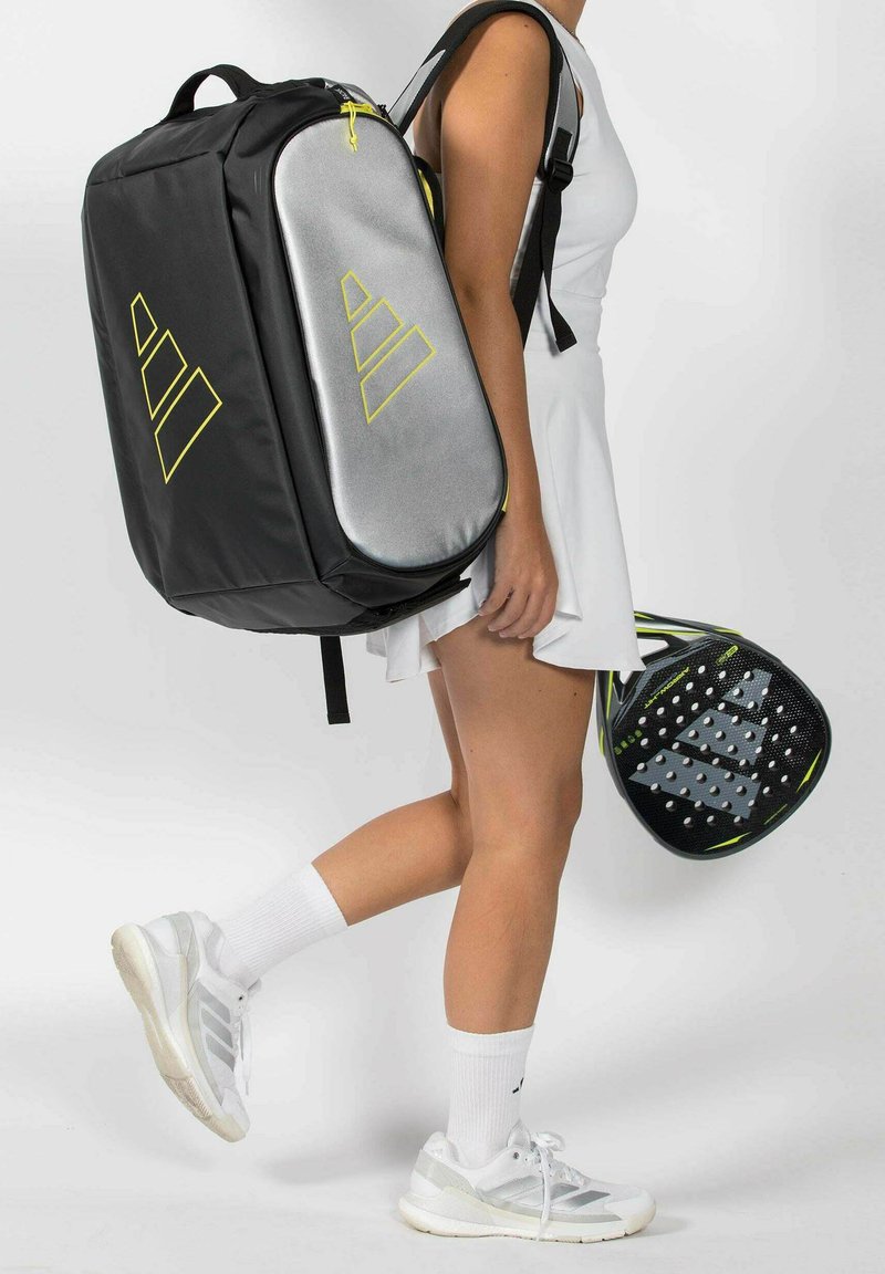 Femme en tenue de sport blanche portant un sac à dos de tennis noir et argent et tenant une raquette de paddle noire et verte, en marchant.