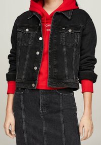 Personne portant un hoodie rouge sous une veste en denim noir et une jupe en denim noir assortie, les mains reposant tranquillement sur les côtés, fond clair.