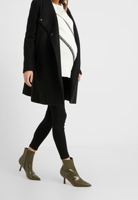 Abrigo de lana negro sobre un suéter blanco con un patrón diagonal negro, acompañado de unos jeans negros ajustados y botines de punta afilada en verde oliva.