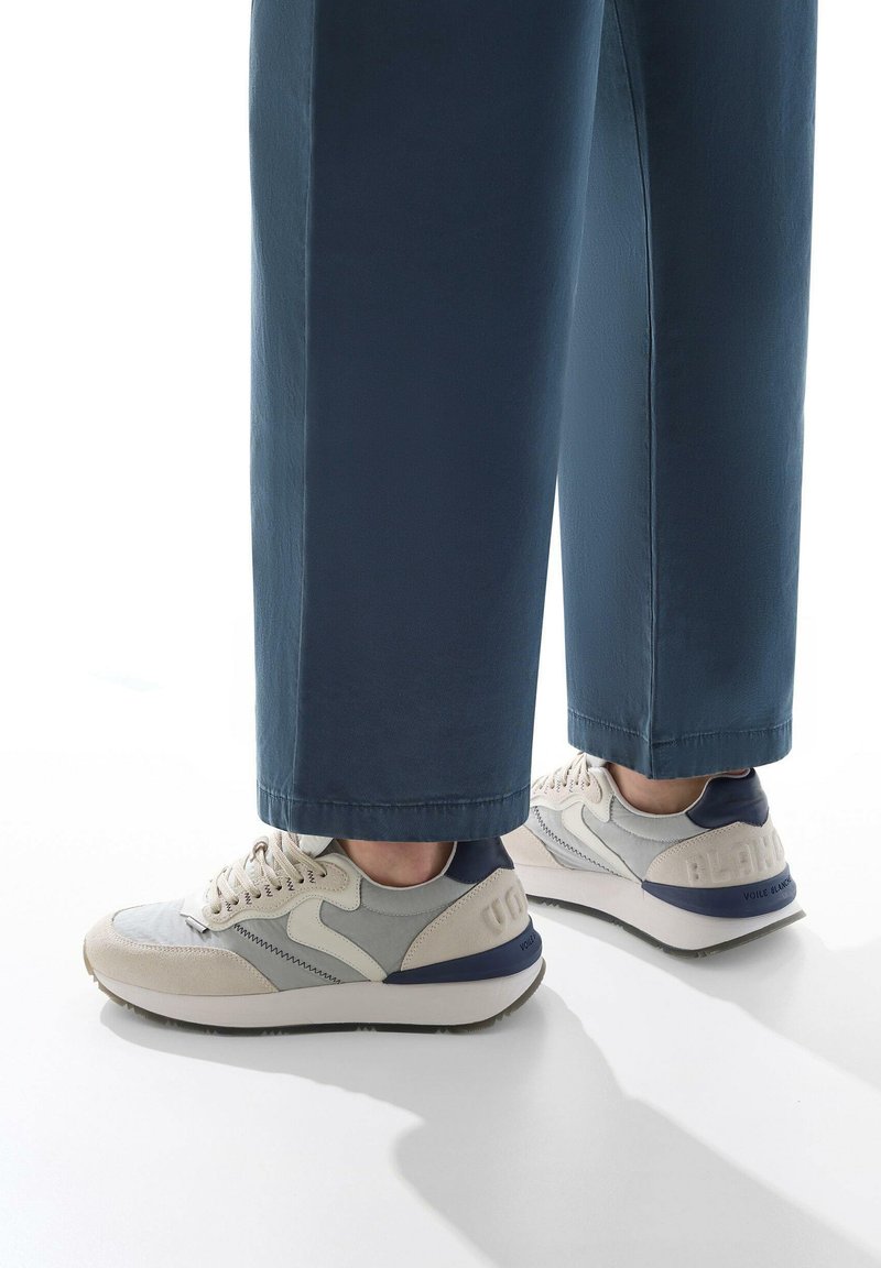 Sneaker con una combinazione di colori beige e grigio chiaro, con un accento blu sul tallone, tessuto texturizzato e suola imbottita, abbinate a pantaloni blu navy.
