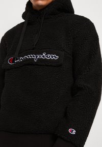 Person trägt einen schwarzen Fleece-Hoodie von Champion, mit sichtbar aufgesticktem Markenlogo auf der Klappe der Brusttasche und am Ärmelbund.
