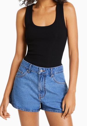Kvinde iført en sort ærmeløs ribstrikket top og højttaljede midtblå denimshorts med forlommer og knaplukning.