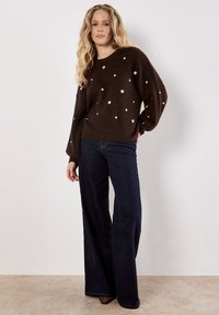 Maglione lavorato a maglia marrone con motivi a pois bianchi, colletto rotondo e maniche a palloncino, abbinato a jeans flare in denim scuro.