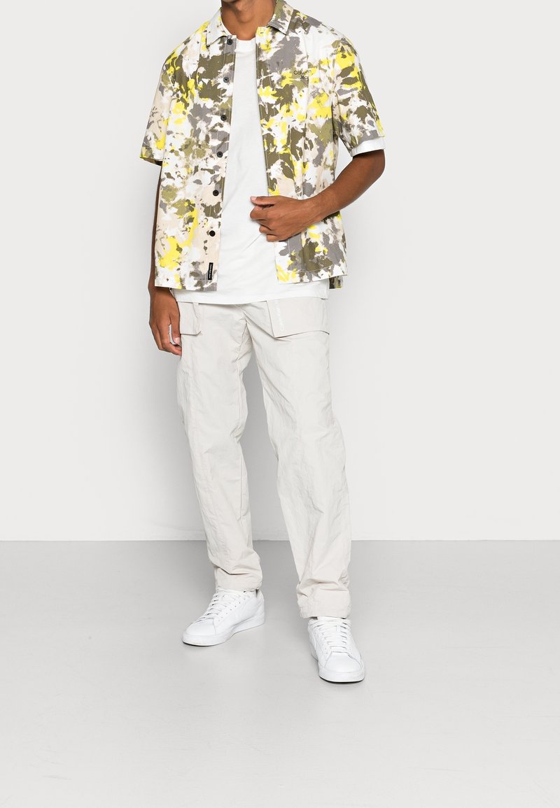 Homme portant des baskets blanches, un pantalon cargo beige clair, un t-shirt blanc et une chemise à manches courtes à motif abstrait jaune, gris et blanc.