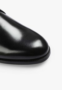 Zapato de vestir de cuero negro con textura pulida y suave, punta redondeada, detalles cosidos y suela plana de goma.