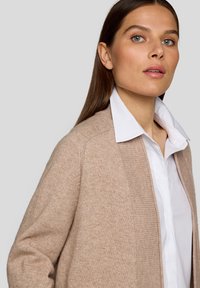Beiger Cardigan, weiches Strickmaterial, offene Vorderseite, gerippte Akzente an Bündchen und Saum, getragen über einem weißen Hemd mit Kragen, minimalistisches Design.