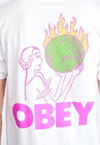 White cotton T-shirt featuring a pink graphic of a woman holding a green globe with a checkered pattern, surrounded by yellow flames, and "OBEY" text.